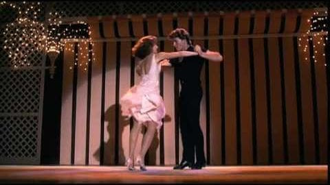 A young adult female dancer in a white ruffled dress performs on an indoor stage with decorative elements, accompanied by a male figure in a dark suit under warm stage lighting.