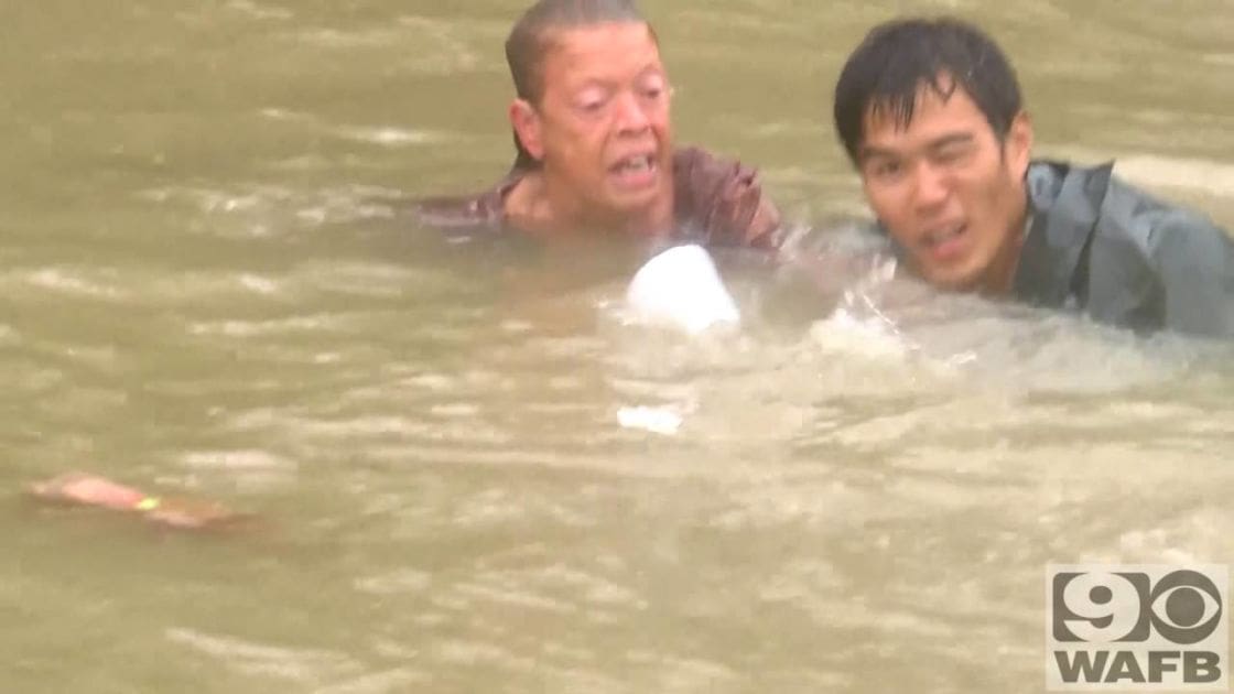 Middle-aged male rescuers in floodwaters assisting each other against murky water and turbulent conditions, likely during daytime.