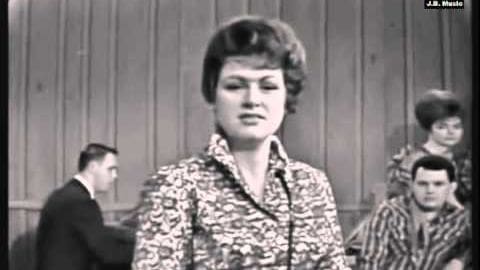 Young adult female wearing a patterned blouse stands in an indoor studio with wooden paneling, surrounded by other individuals dressed formally and casually.