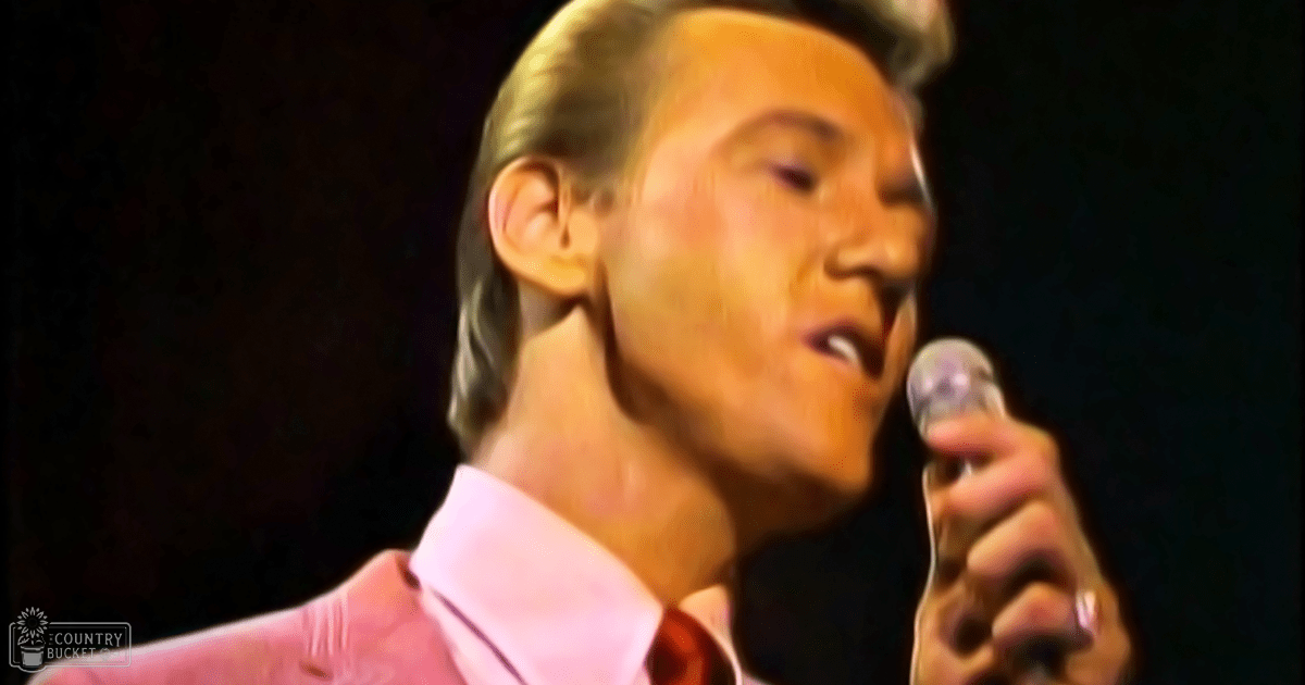 An adult male singer wearing a formal pink shirt and red tie, performing into a microphone at an indoor concert or event setting with focused stage lighting.