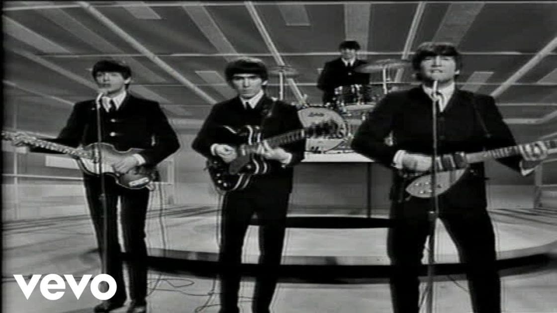 A 1960s-style musical performance in a professional recording studio with young adult male musicians wearing black double-breasted suits, playing guitars and singing into microphones.