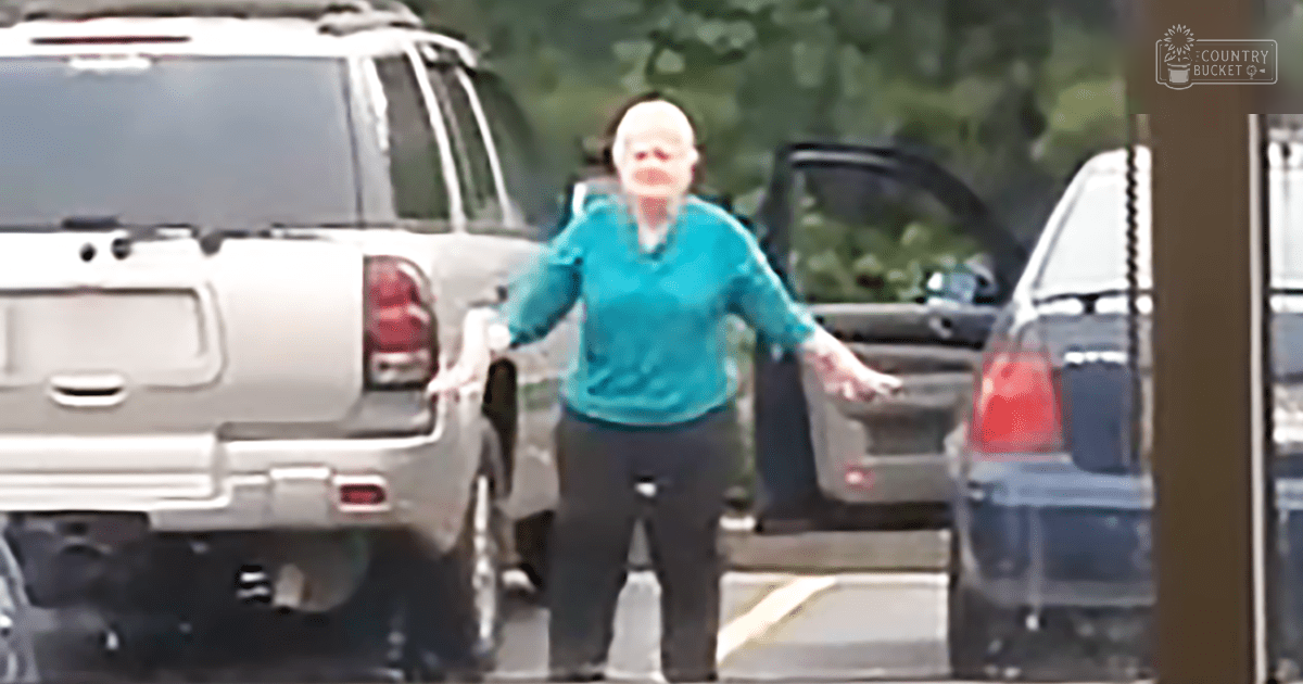 An older adult female in teal sweater and dark pants stands in an outdoor parking lot, late spring or summer day with natural lighting.