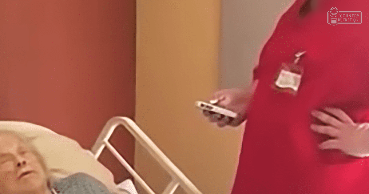 An adult female healthcare professional wearing red scrubs with a name tag and badge holder, holding a smartphone in both hands next to an older adult woman in a hospital gown or bedsheet with a floral pattern. The setting is a hospital room with warm-toned walls and furniture indicating a medical environment.