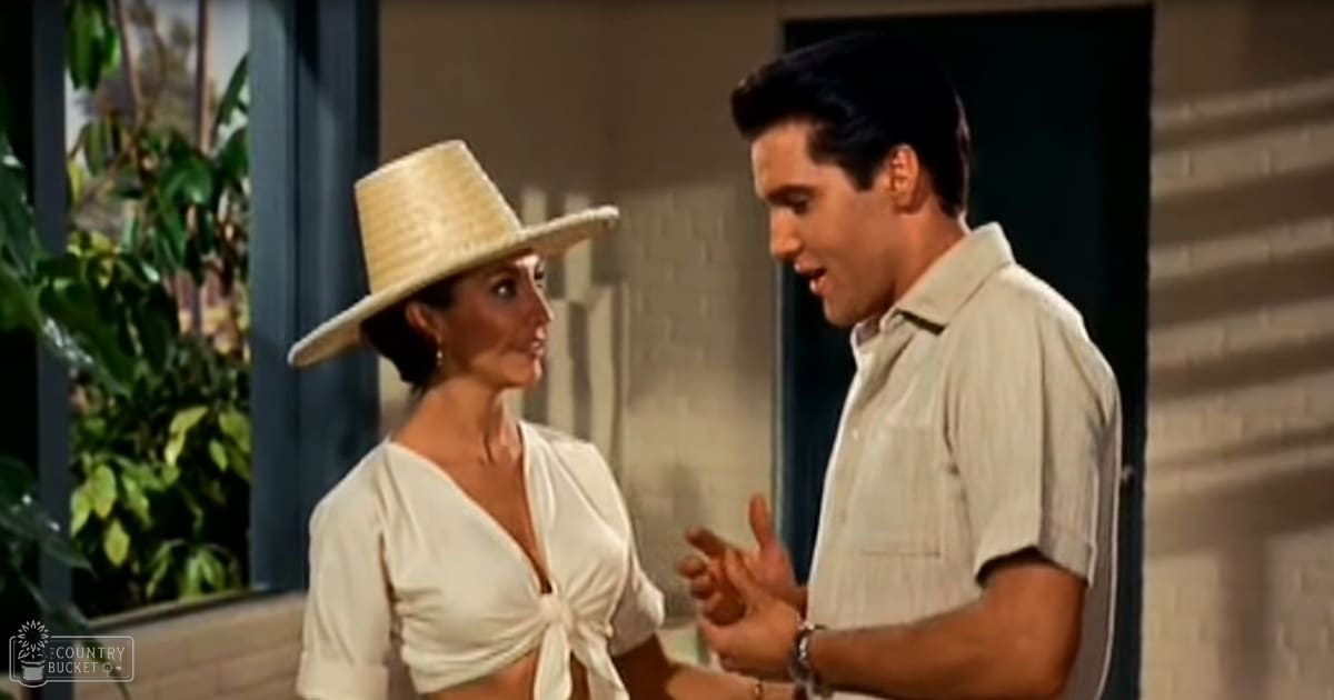 An adult male singer in a young adult age band wearing a light-colored button-up shirt with short sleeves and a watch on his left wrist converses with an adult female middle-aged or older, wearing a wide-brimmed straw hat, a white blouse with a deep V-neck and tied front, short sleeves, and earrings. The setting is outdoors near a building during the daytime in spring or summer.