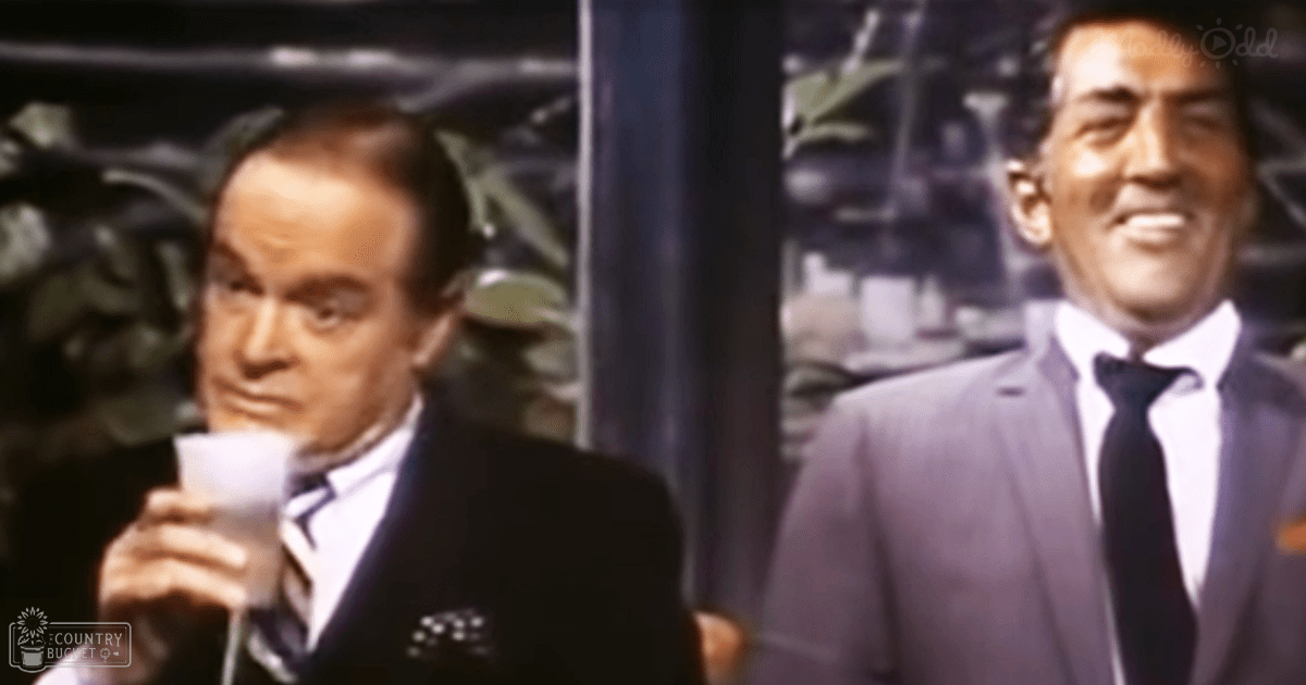 An indoor scene from a classic film or television show with two middle-aged men in well-fitted suits, set against an evening ambiance with greenery visible in the background.