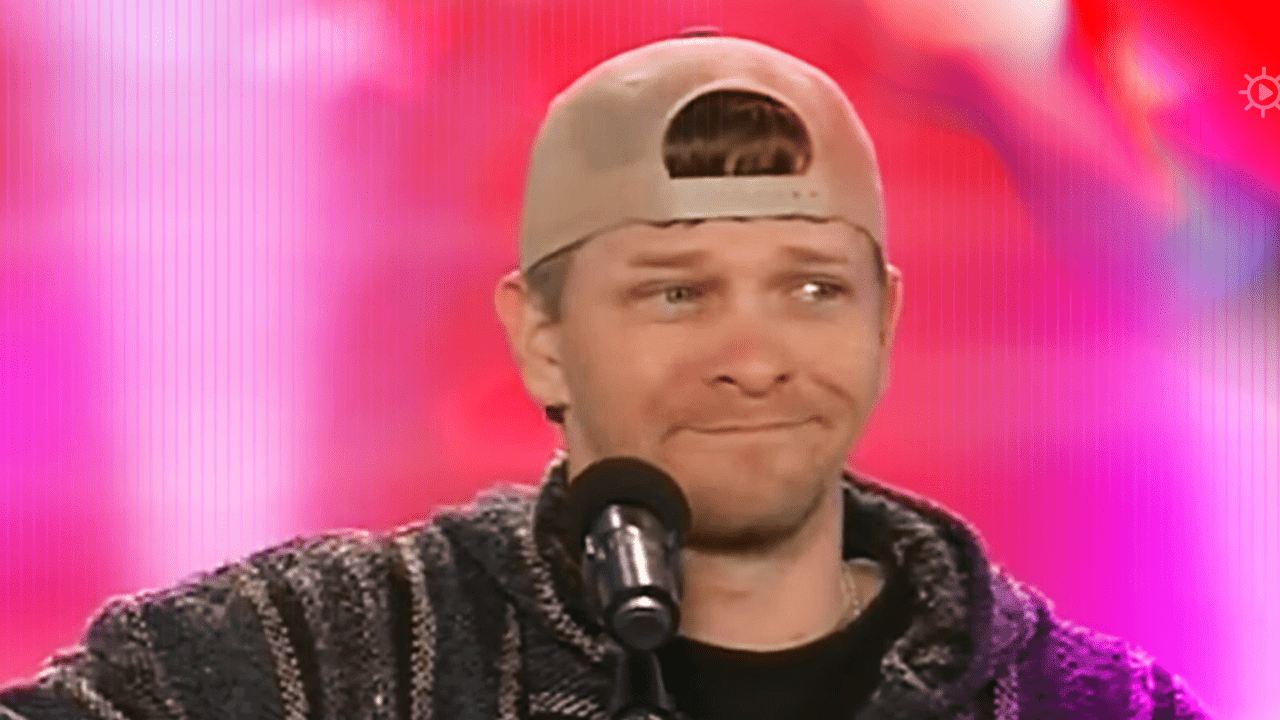 A young adult male wearing a patterned hoodie and baseball cap, standing in an indoor stage setting with vibrant pink and red lighting effects.