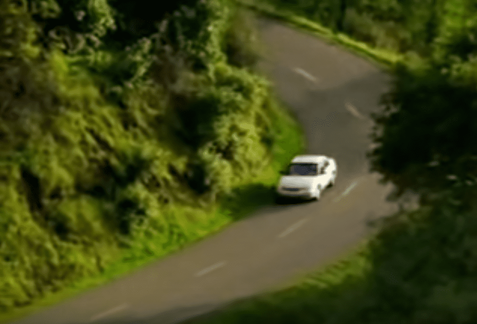 A white van travels on a curving rural road surrounded by lush greenery, suggesting late afternoon light with shadows cast by trees.