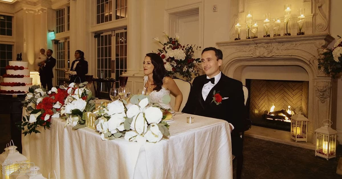 A couple at their wedding reception in an elegant indoor setting with a large multi-tiered wedding cake, wine glasses, and floral arrangements.