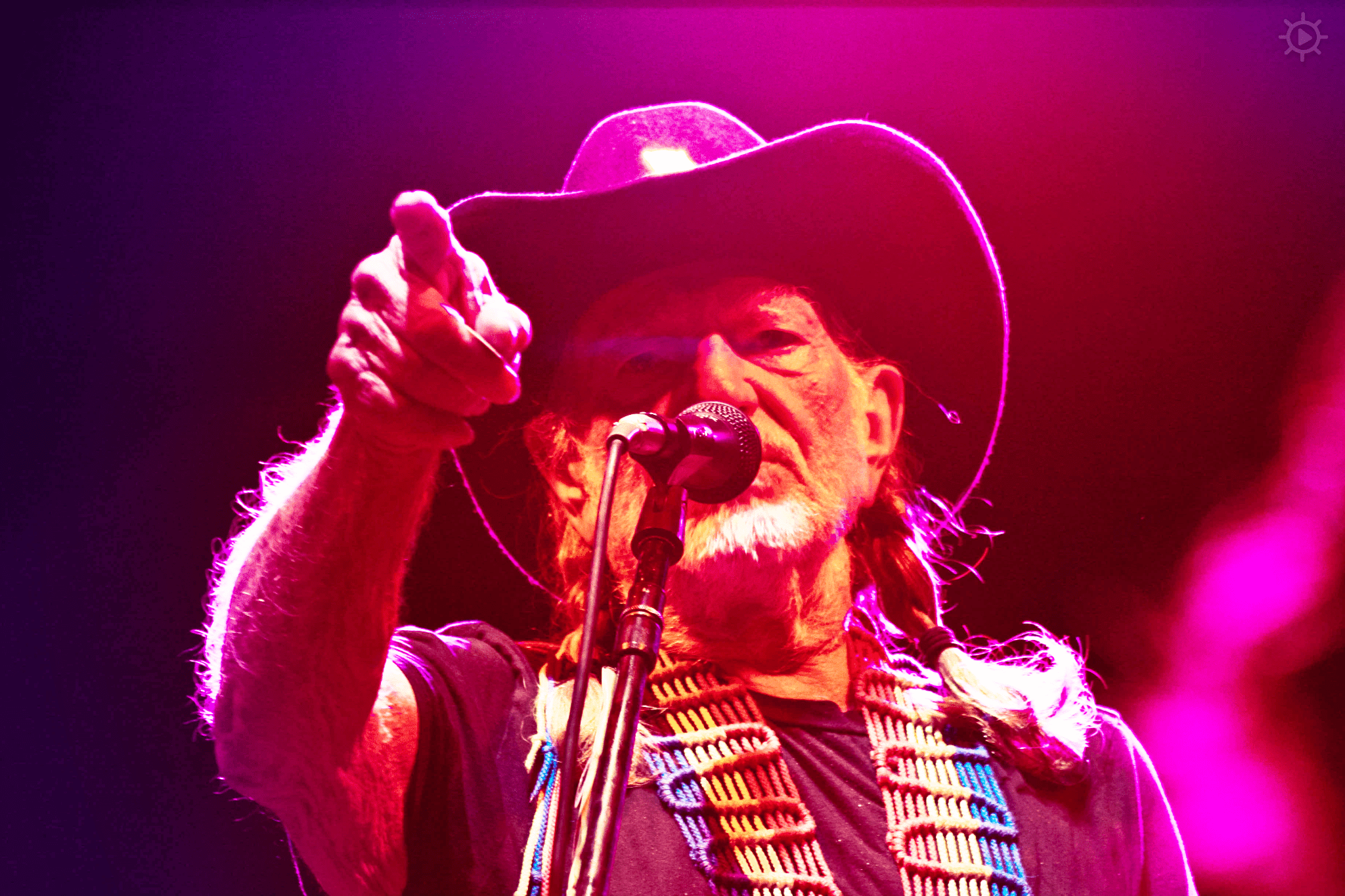 A middle-aged man with dreadlocks, wearing a black t-shirt with colorful trim and a wide-brimmed cowboy hat, performs at an indoor concert venue under vibrant red and purple stage lighting.