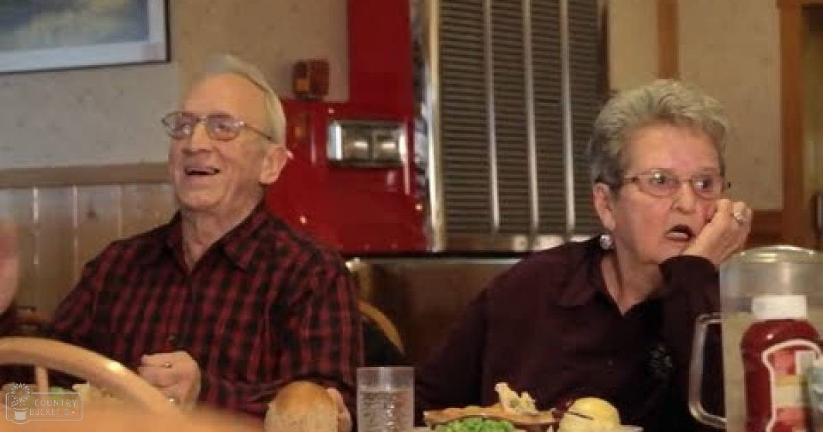 An older adult male and female wearing casual attire with red plaid shirt, dark maroon shirt, dining at a diner-style restaurant setting with food on the table.