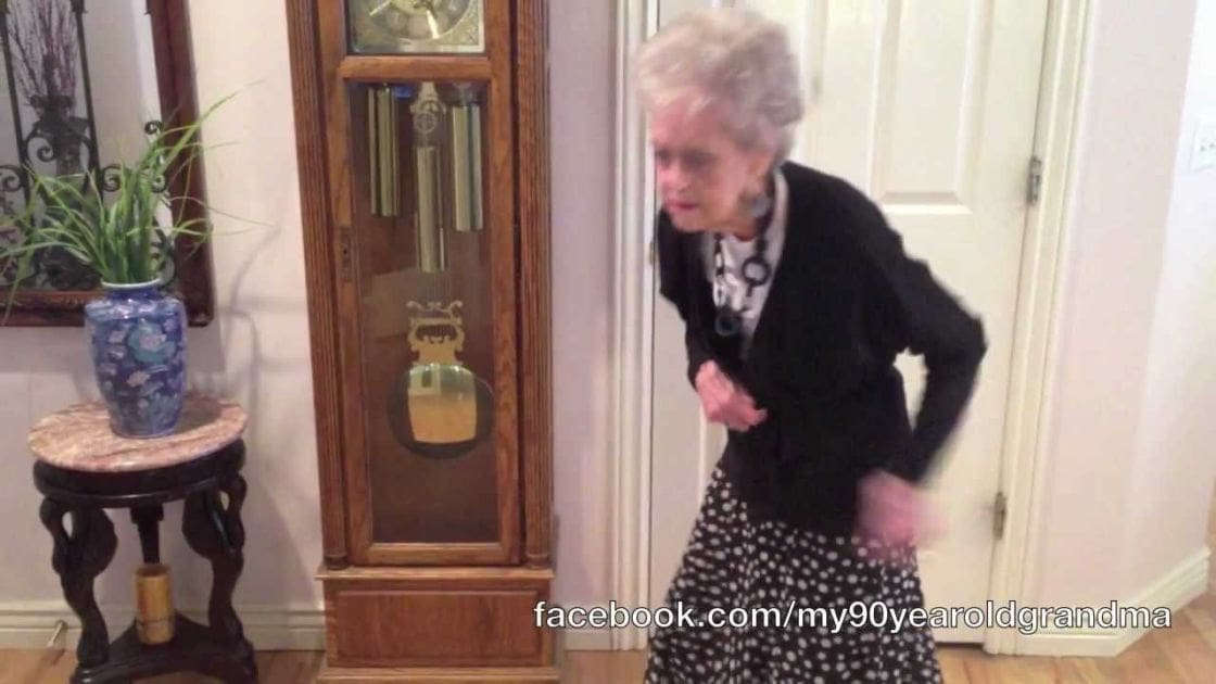 An older adult woman in a home setting wearing a black blazer, patterned skirt, white blouse, and necklace. The scene includes a grandfather clock, marble table with blue vase, and soft lighting.