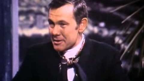 A middle-aged adult male wearing a dark-colored suit jacket with a high collar, white collared shirt, and bow tie in an indoor setting.