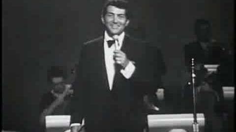 Adult male performer in a formal black tuxedo with white shirt and bow tie, seated on stage under dim lighting.