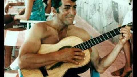 A middle-aged man with short dark hair plays a guitar in an outdoor setting with white chairs and tables.