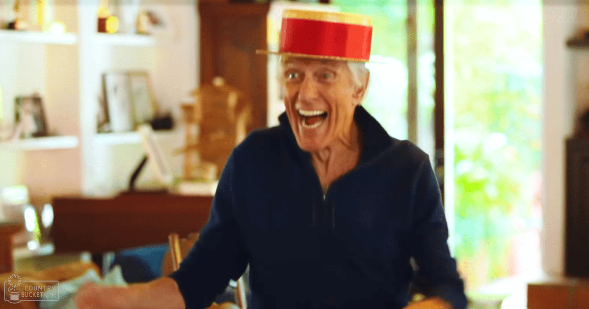 An older adult male in a relaxed home setting, wearing a dark blue zip-up sweater with long sleeves, indoors under natural light. Shelves hold various items, wooden furniture is visible, and greenery outside suggests spring or summer.