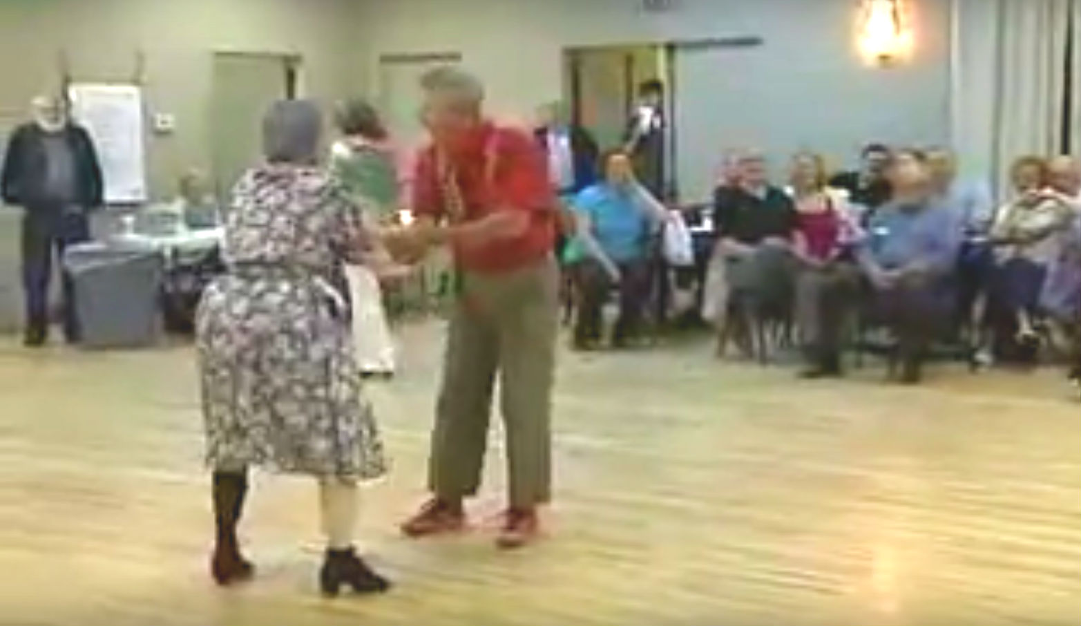 An older couple dancing in a community center with other spectators seated around them.