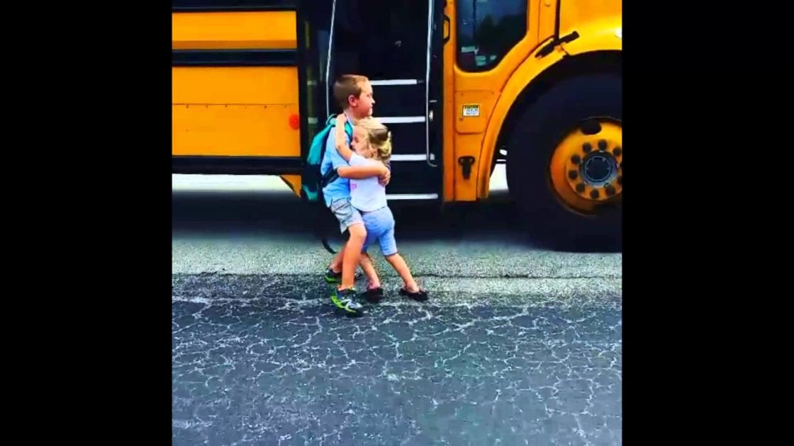 Two young children near a yellow school bus at what appears to be a school drop-off or pick-up point. The scene is outdoors on paved ground with natural daylight.