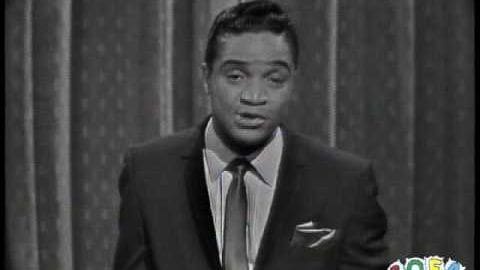 A man wearing a dark suit jacket with a white pocket square stands indoors against a patterned curtain backdrop in black and white.