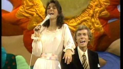 Young adult female singer in a white ruffled dress with a belt, performing into a microphone alongside another young adult male seated in a dark suit jacket and striped tie on an ornate stage with colorful floral drapes.