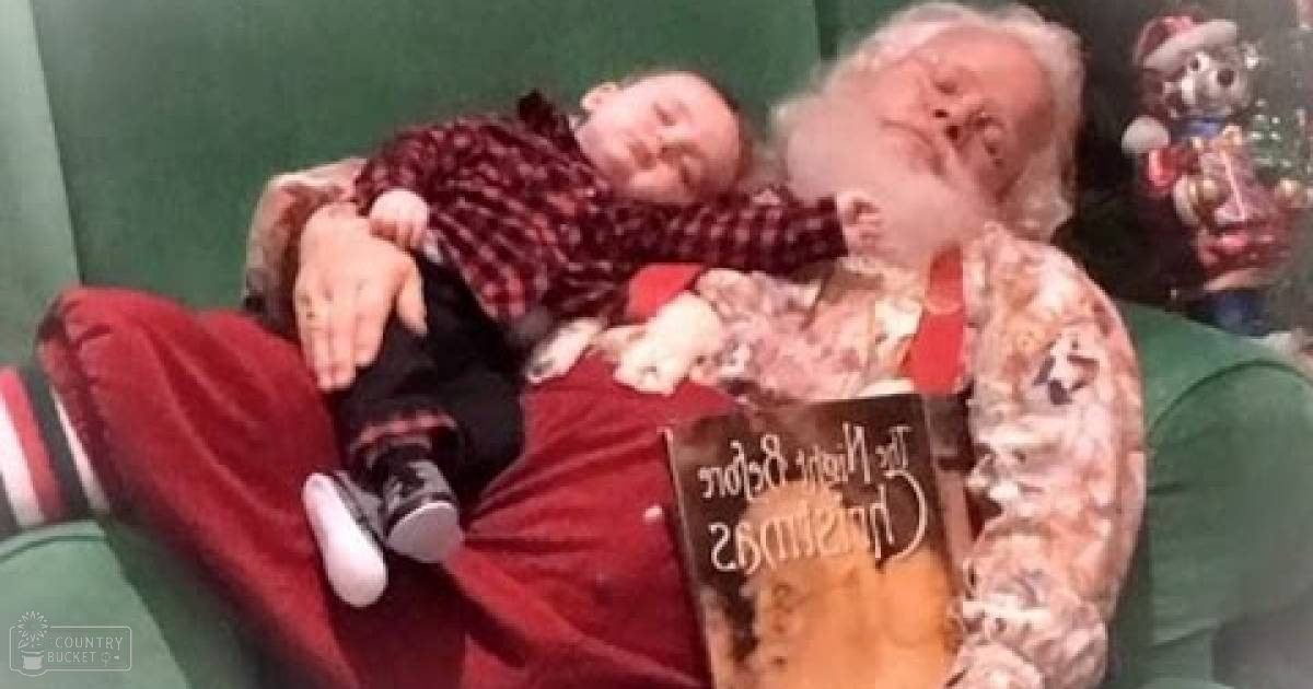 A toddler girl in a red and black checkered shirt sleeps on Santa Claus’s lap amidst Christmas-themed decorations indoors.