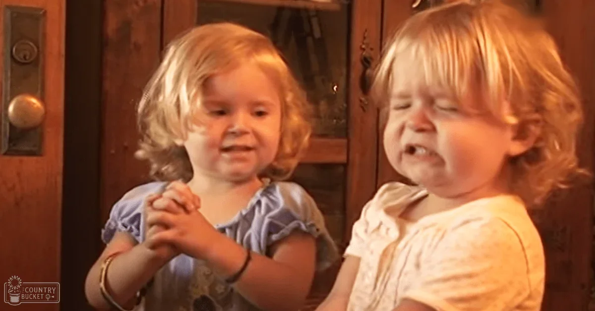 Two toddlers in a home setting with natural lighting, wearing light blue and white dresses near a wooden door and dark cabinet.