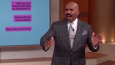 An adult male in a tailored gray plaid suit jacket with a pocket square and lapel pin, standing against the backdrop of a television studio set displaying text messages.
