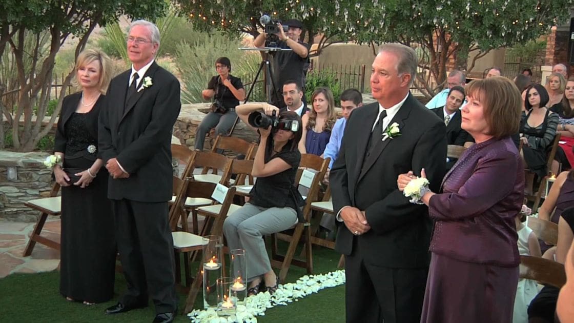 A wedding ceremony unfolds outdoors in a garden setting with wooden folding chairs and greenery surrounding the area. Two adults on each side wear formal attire, while another sits in casual clothes taking photos.