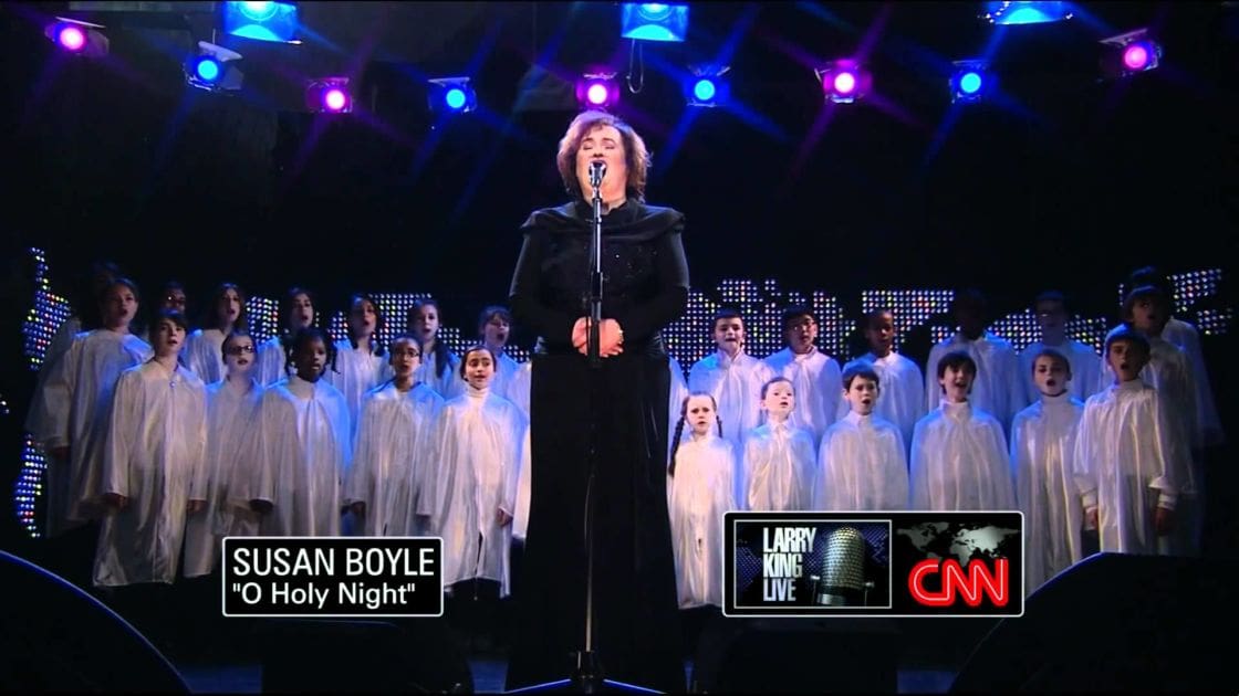 Susan Boyle performing 'O Holy Night' at a CNN event hosted by Larry King Live, with choir members wearing white robes and stage lights in purple and blue hues.