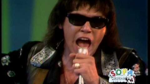 An adult male singer in a black leather jacket with patches, holding a microphone and wearing sunglasses. The setting appears to be a studio or stage setup for a live performance.