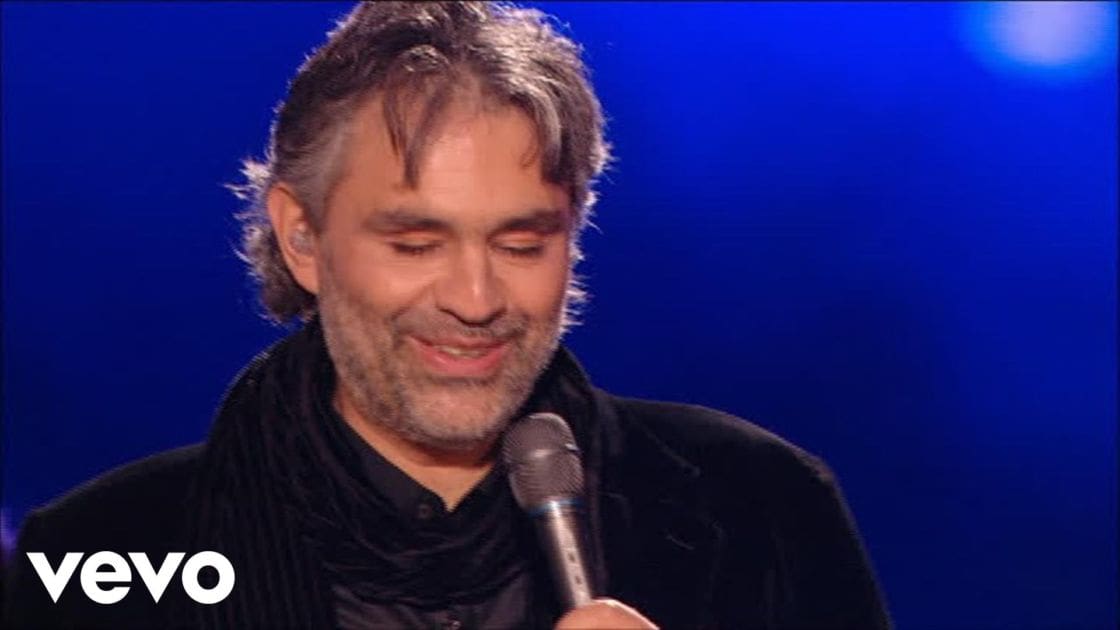 A middle-aged adult male in a relaxed fit black shirt with a high collar, holding a microphone on an indoor stage or studio set under artificial blue light effects and spotlighting.