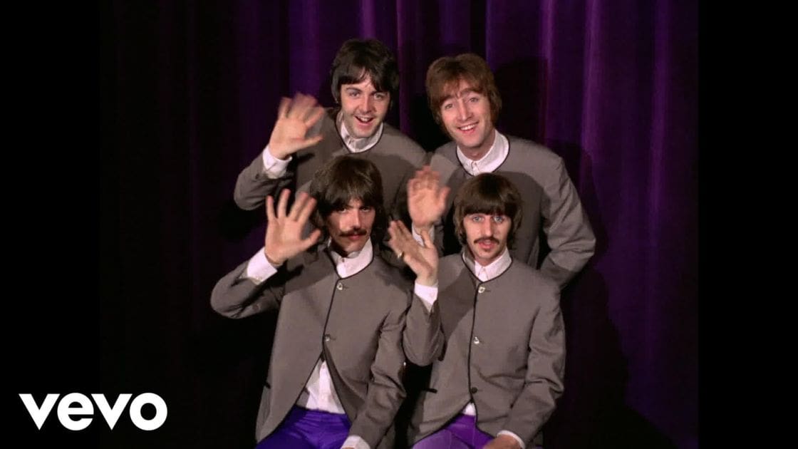 Four young adult males in light gray jackets with rolled-up sleeves, standing against a solid purple curtain indoors, possibly on stage or at an event.