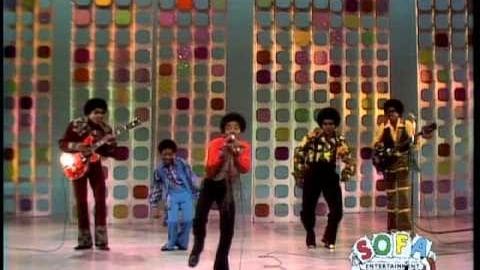 A musical performance group on stage with a colorful backdrop in an indoor venue likely from the 1970s.