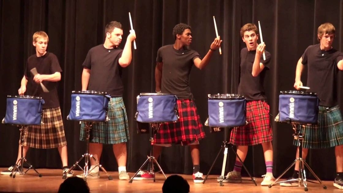 Teenagers performing a drumming group on stage with simple curtains, daytime lighting, in a school or community hall setting.