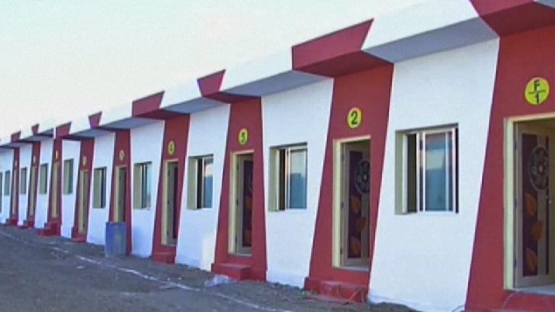 A row of small identical houses numbered from 1 to 9, each with a red and white exterior, triangular roof section, and windows on either side of the door. Decorative leaf patterns adorn the doors.