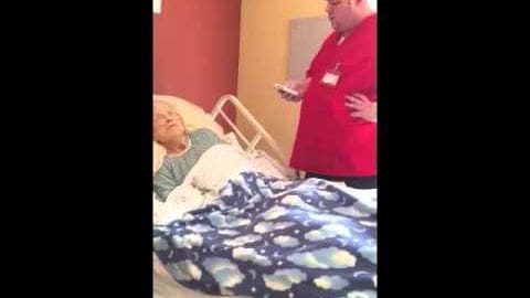 Indoor healthcare setting, likely a hospital room; adult male standing in red scrubs with name tag, older adult lying on bed wearing light green patterned shirt and dark pants.