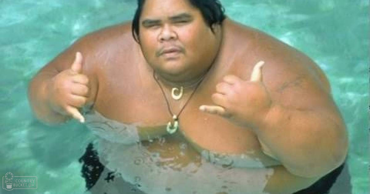 Adult male in dark swim trunks at an outdoor swimming area, wearing a circular pendant necklace under natural daylight.