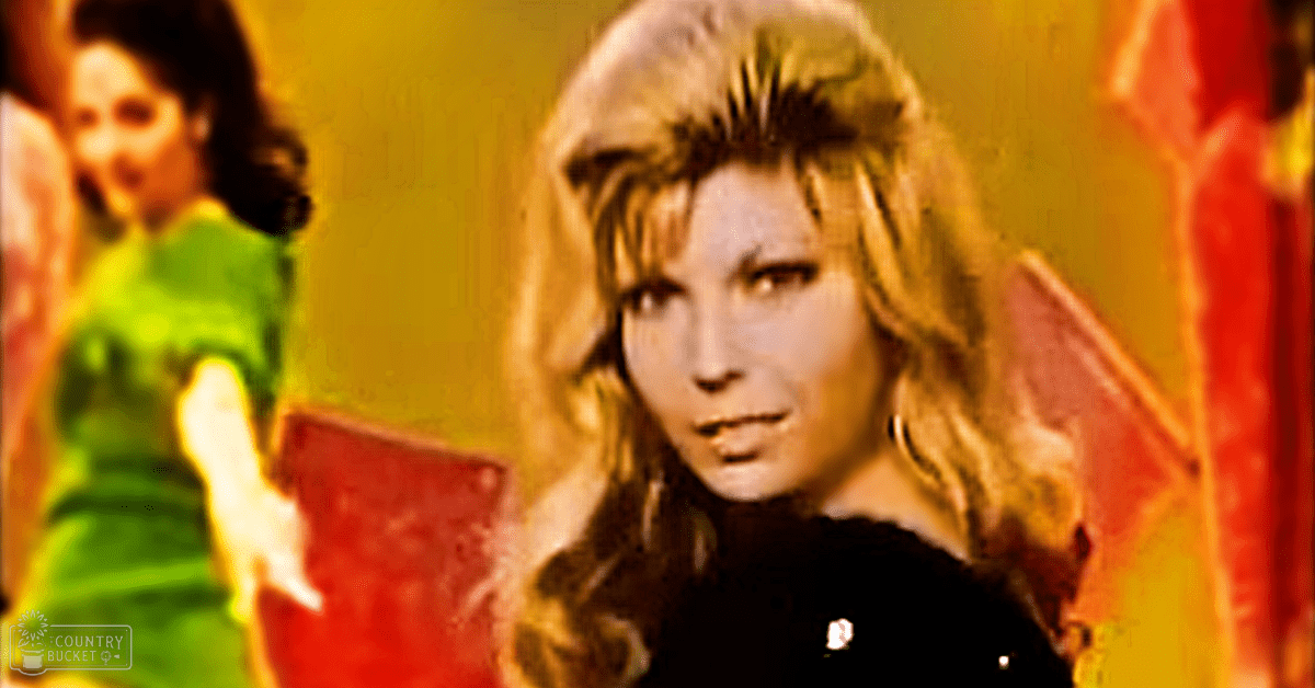 An adult female with long, wavy blonde hair wearing a black garment with a high collar and a white logo, large hoop earrings. A young girl in a bright green dress with short sleeves and white gloves stands nearby.
