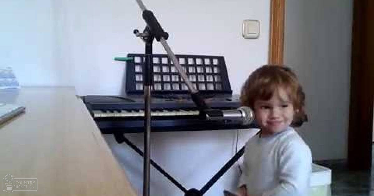 A toddler in a light-colored long-sleeve shirt indoors, near sheet music and a microphone, suggesting a musical setting.