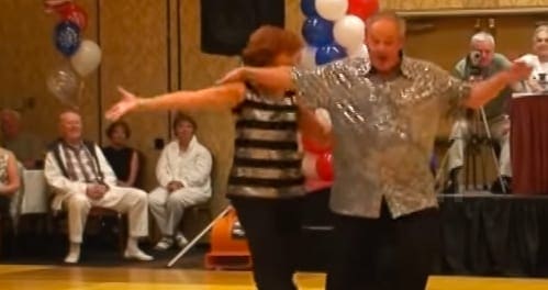 A community dance performance in an indoor venue with a stage, red and white balloons, and audience watching. Two adults are dancing; one is wearing a black and white striped top and dark pants, the other in a patterned beige shirt and black pants.