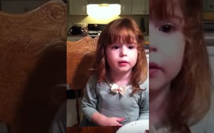 A toddler girl wearing a light green long-sleeved shirt with a small bow at the collar stands in an indoor kitchen setting, surrounded by wooden furniture and home decor items.