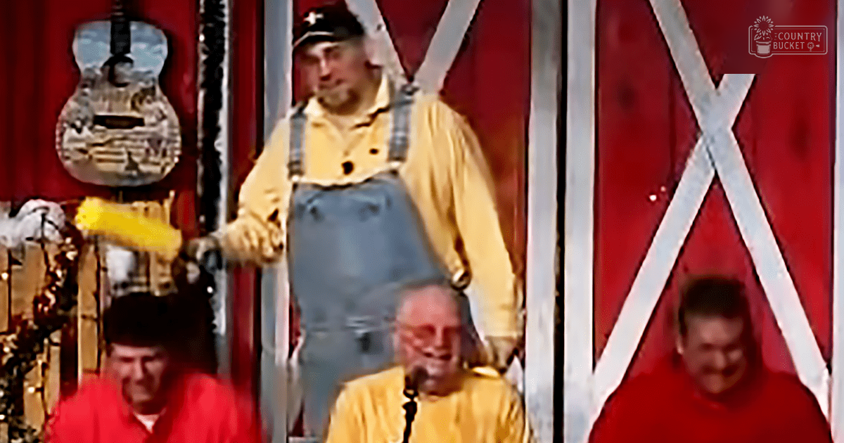 Middle-aged adult male performer in a barn-themed setting, possibly at a country music event; red barn door with white X's and festive decorations suggest holiday season (Christmas lights)