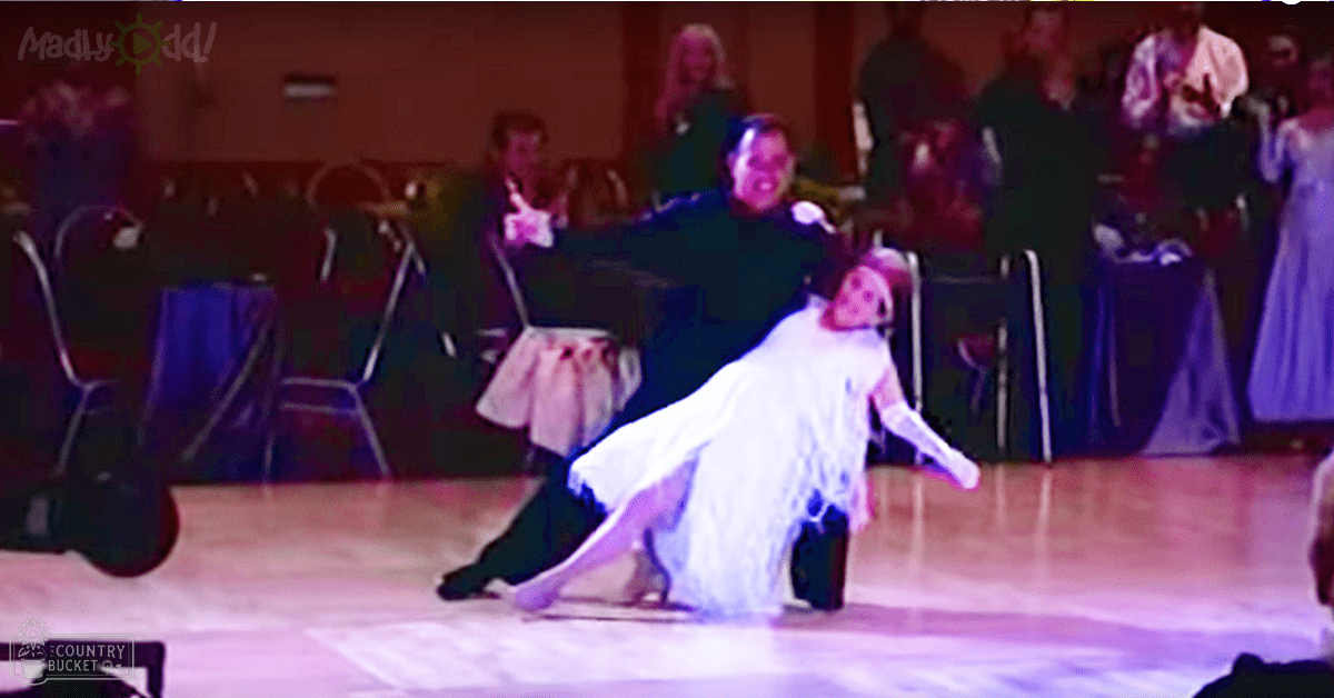 A couple dancing in a ballroom setting, with a man wearing a dark tuxedo and a woman in a white ball gown, surrounded by guests clapping under evening lighting.