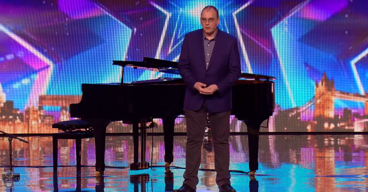 A middle-aged adult male stands on stage at a talent show or performance setting, wearing a dark blue blazer over a patterned shirt and gray trousers.