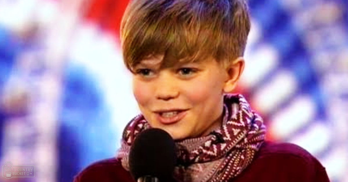 A young child wearing a maroon sweater with a patterned scarf stands on what appears to be an indoor stage during a public event, surrounded by blurred audience lights.
