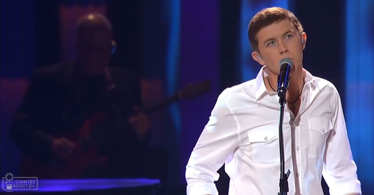 A young adult male performer wearing a white button-up shirt stands on stage, accompanied by an adult male band member holding a guitar.