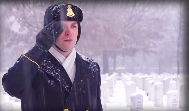 A young adult male in a cemetery during winter, wearing a dark-colored coat with a high collar and fur-lined hood. The setting is an outdoor cemetery with snow-covered tombstones and trees.