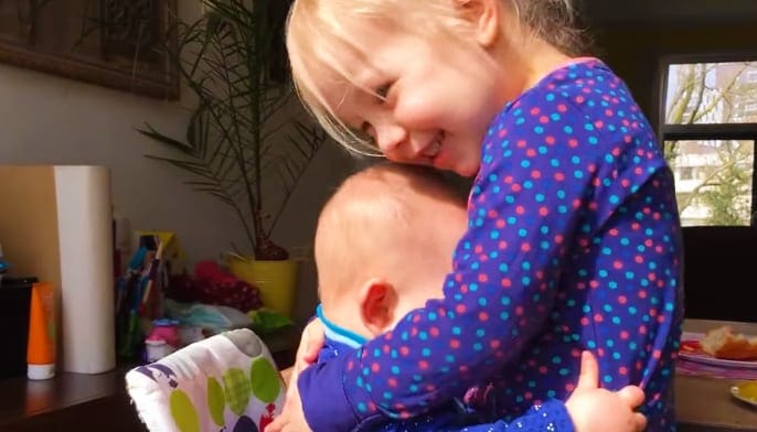 A young child holding an infant in a home setting with natural daylight illuminating the scene.