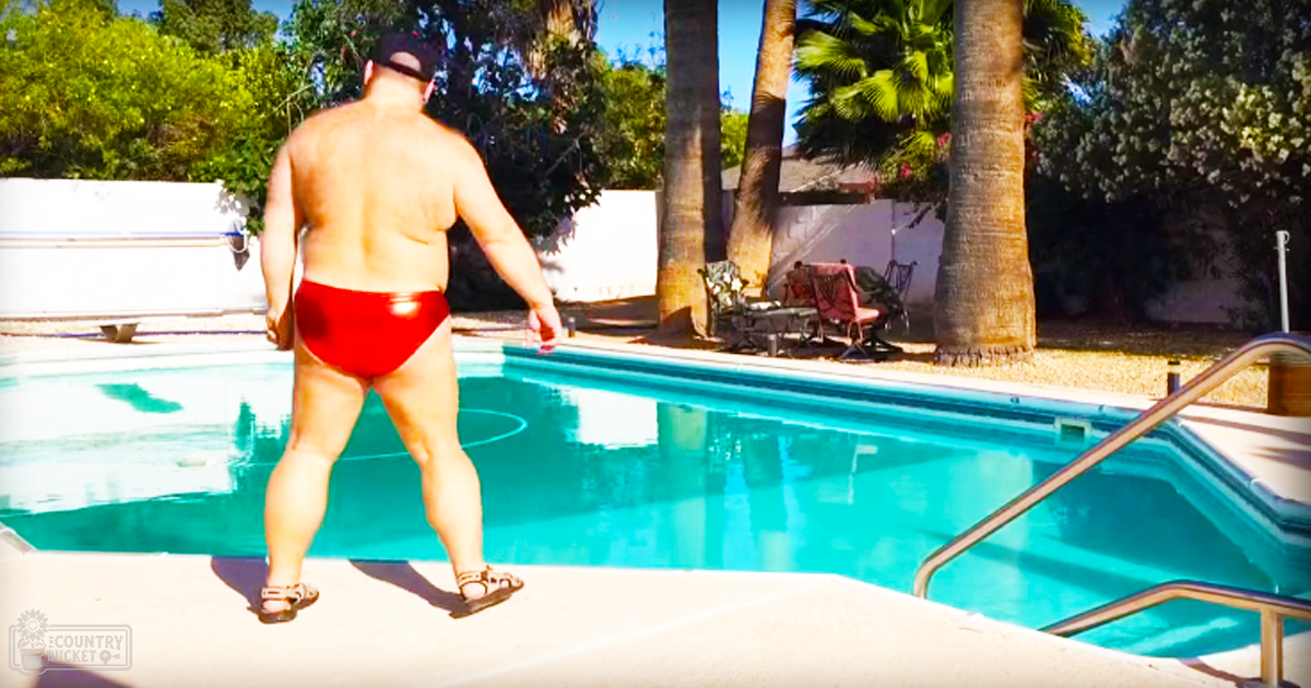 Adult male wearing bright red swim trunks with shiny synthetic material, standing by a swimming pool surrounded by palm trees and outdoor furniture under bright daylight at what appears to be summer or late spring.