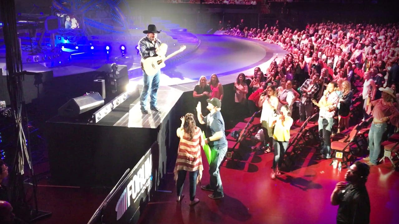 A male performer in a black cowboy hat and denim jeans plays guitar on stage at an indoor concert venue.