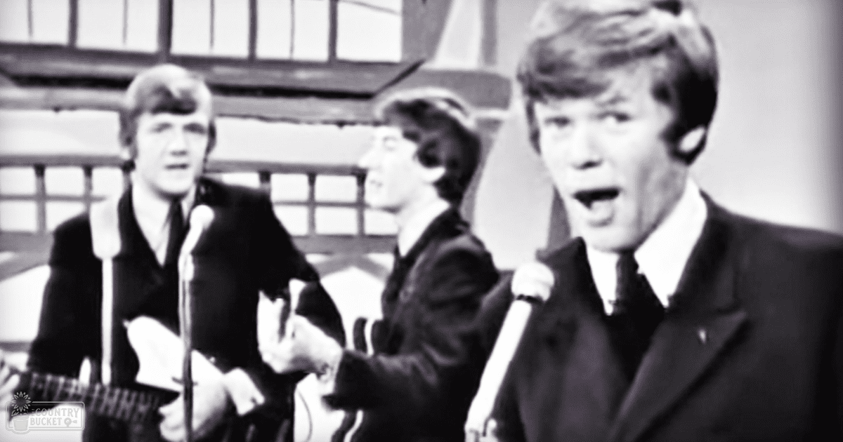 A musical performance scene from the 1960s with young adult males in formal suits holding guitars and microphones, set indoors on a stage-like structure with wooden beams visible in the background.